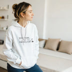 Female model wearing a white hoodie with the black “You Were Never Ordinary” statement design, standing in a bright minimalist living room with hands in the front pocket.