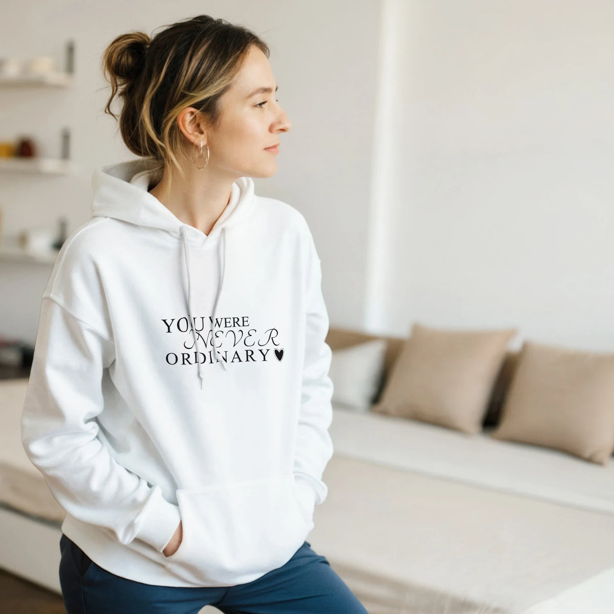 Female model wearing a white hoodie with the black “You Were Never Ordinary” statement design, standing in a bright minimalist living room with hands in the front pocket.