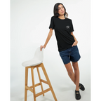 Female model wearing a black “Color the Grey” unisex T-shirt with a subtle white PK monogram on the front, styled with shorts.