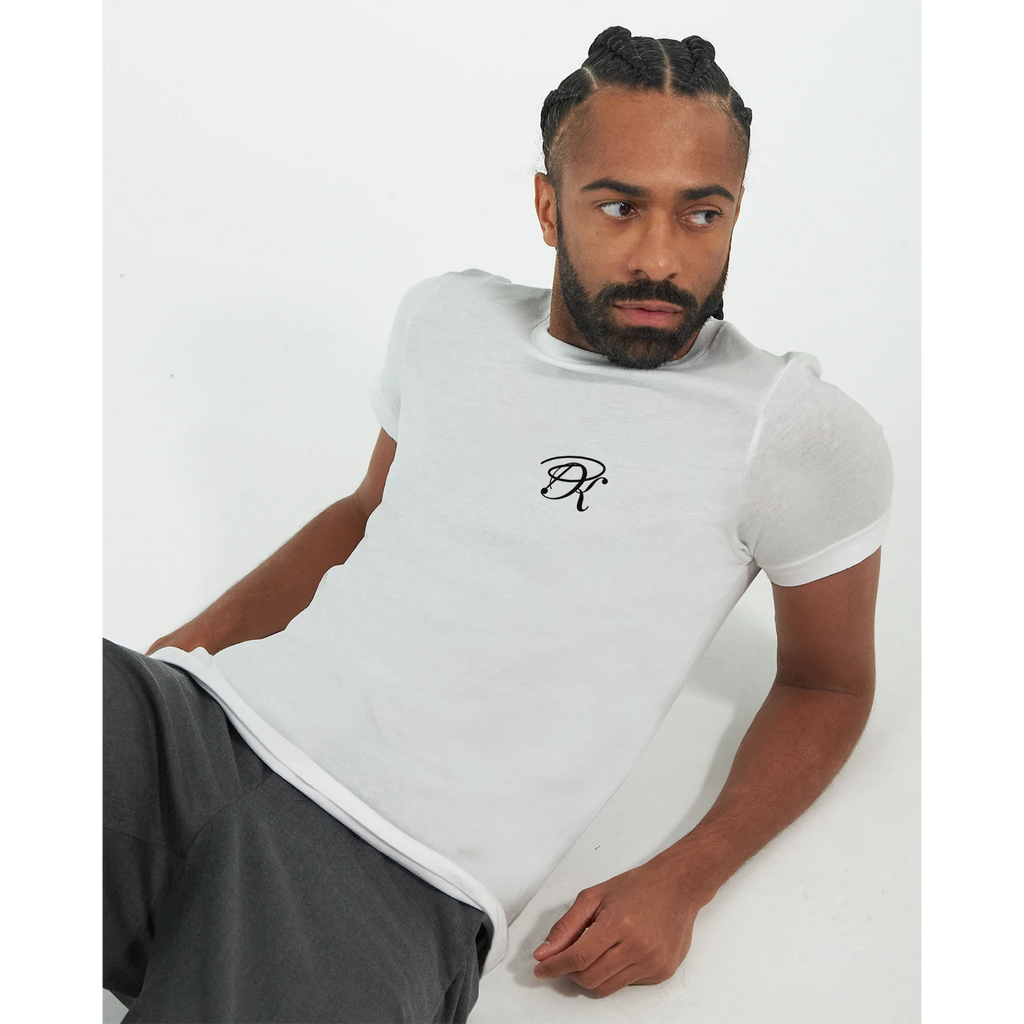 Male model wearing a white T-shirt with a small black PK logo on the chest, styled with dark trousers in a clean, minimal studio setting.