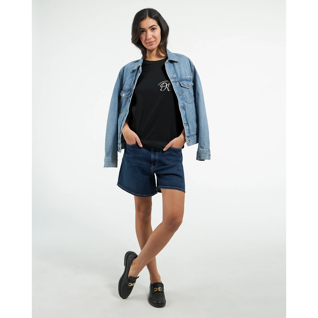 Female model wearing a black unisex statement T-shirt with a small white PK monogram on the chest, styled with a denim jacket and shorts for a casual streetwear look.