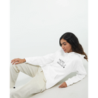 Female model reclining in a white hoodie with the black “You Were Never Ordinary” statement design on the front, styled with light trousers against a clean white studio background.