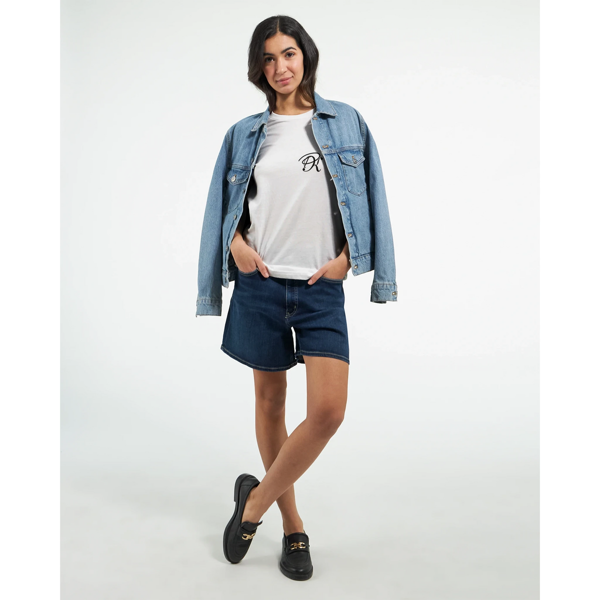 Female model wearing a white T-shirt with a small black PK logo on the chest, layered with a blue denim jacket and paired with denim shorts and black shoes for a casual, modern streetwear look.