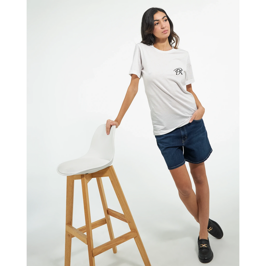 Female model wearing a white T-shirt with a small black PK logo on the chest, paired with denim shorts and black shoes in a clean studio setting for a casual unisex streetwear look.
