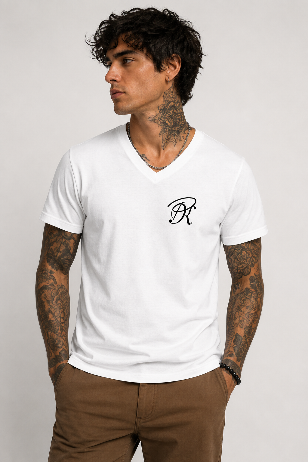 Front view of a tattooed male model wearing a white unisex V-neck T-shirt with a small black PK monogram on the left chest, paired with brown trousers for a clean modern look.