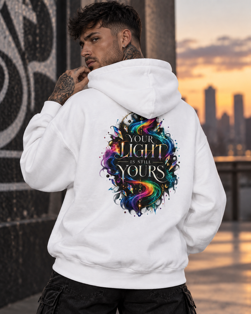 Tattooed men wearing a white hoodie featuring the colorful “Your Light Is Still Yours” back print, photographed in an urban setting at sunset.