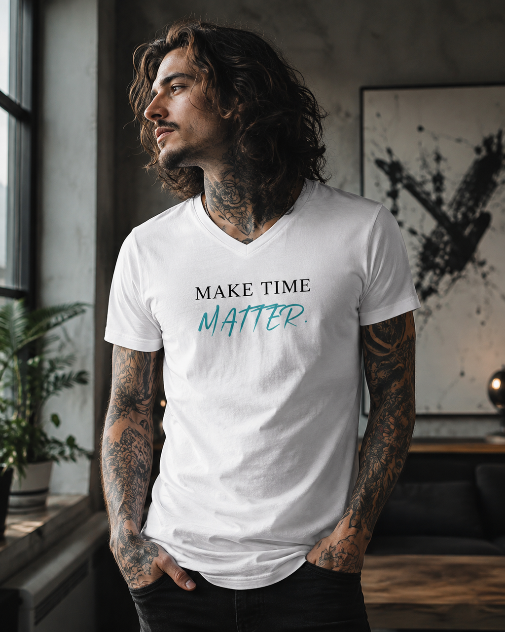 Tattooed long-haired man wearing a white premium unisex V-neck T-shirt with the minimalist motivational statement 'MAKE TIME MATTER.' in black and turquoise lettering. The lifestyle mockup is set in a stylish industrial loft with natural window light, modern artwork, indoor plants, and a contemporary interior, highlighting the clean, inspirational design.