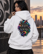 Tattooed woman with long black hair wearing a white hoodie featuring the colorful “Your Light Is Still Yours” back print, photographed in an urban setting at sunset.