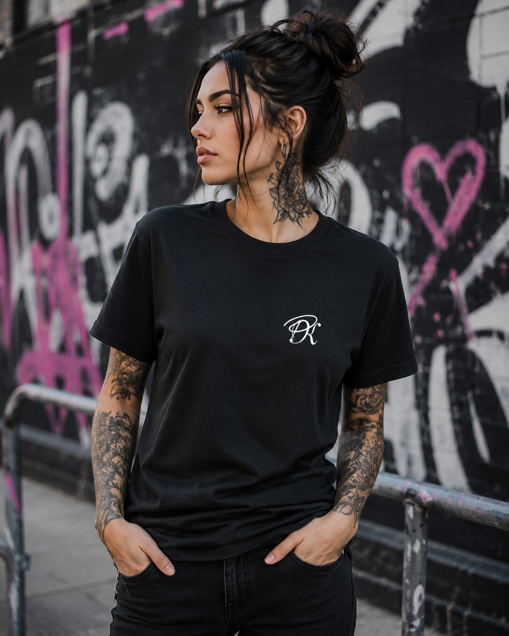 Tattooed female model wearing a black unisex T-shirt with a small white PK monogram on the chest, styled in an urban graffiti setting.