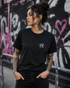 Tattooed female model wearing a black unisex T-shirt with a small white PK monogram on the chest, styled in an urban graffiti setting.
