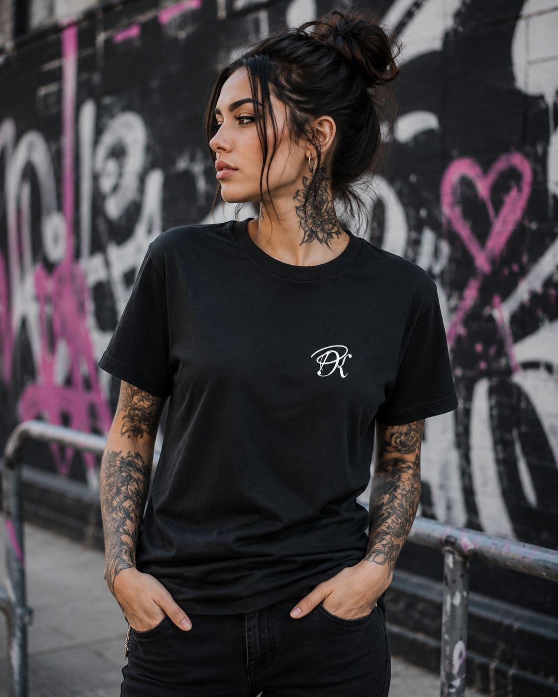 Tattooed female model wearing a black unisex T-shirt with a small white PK monogram on the chest, styled in an urban graffiti setting.